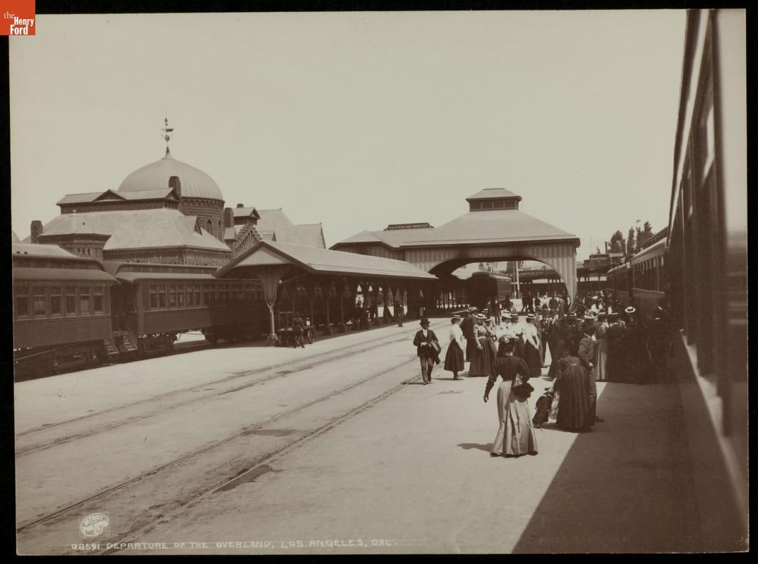 Departure of the Overland, Los Angeles, California, circa 1895 Crowd of people stands by railway tracks with trains on them and a building on the other side of the tracks