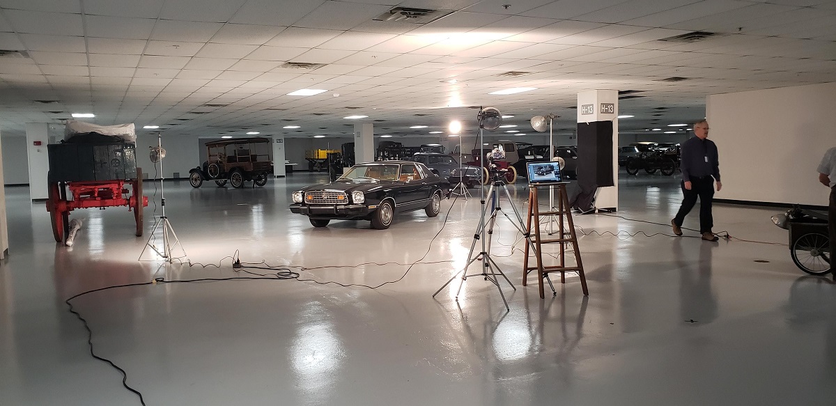 Black car surrounded by other vehicles in warehouse space; lights and camera on tripod pointed at it