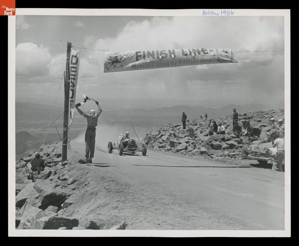 Bobby Unser Crossing the Finish Line, Winner of the 1956 Pikes Peak Auto Hill Climb Race Car approaching banner marked "Finish Line" on dirt road with rocks on either side and steep dropoff on one side, mountains visible in background