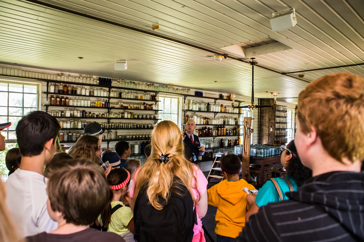 Menlo Park Laboratory in Greenfield Village A group of children watch a man talking in a room filled with bottles and jars on shelves on the wall