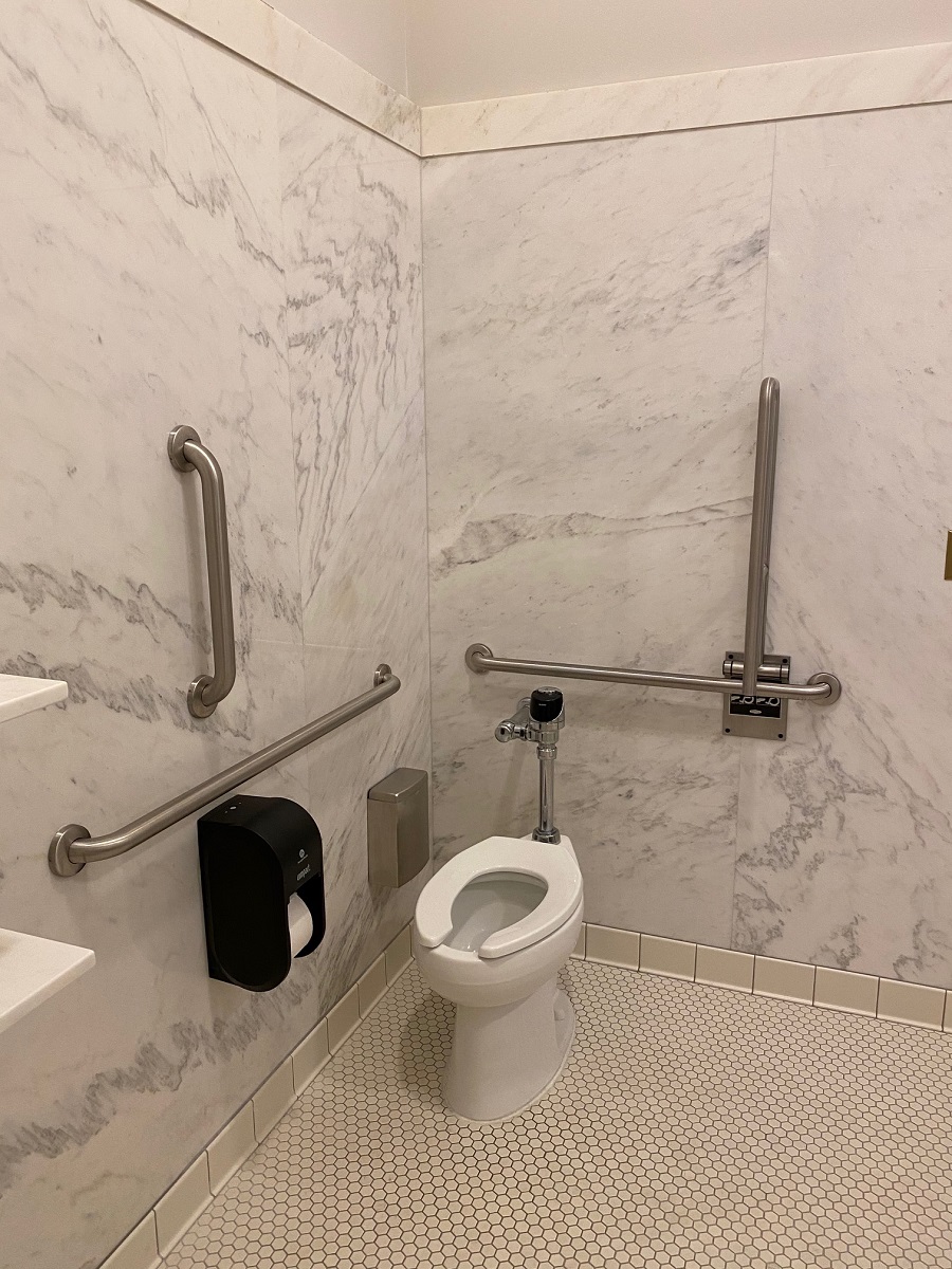Toilet surrounded by vertical and horizontal grab bars in space with tile floor and marble walls