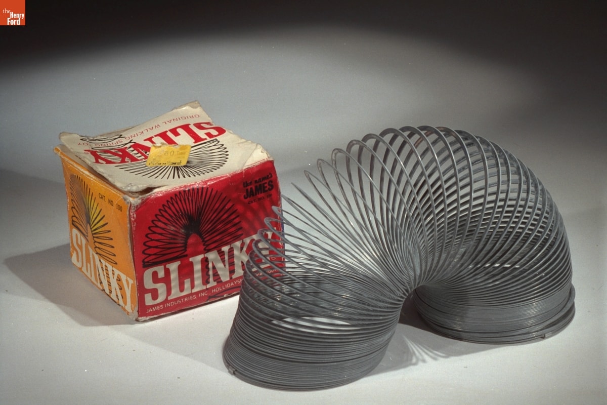 Image with metal coil (Slinky toy) in arch shape in front of yellow and red with text and image of Slinky coil