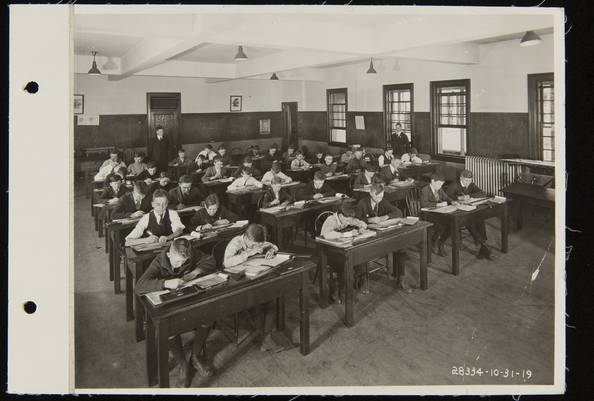 Group of students work at desks in a classroom with a teacher standing in the back