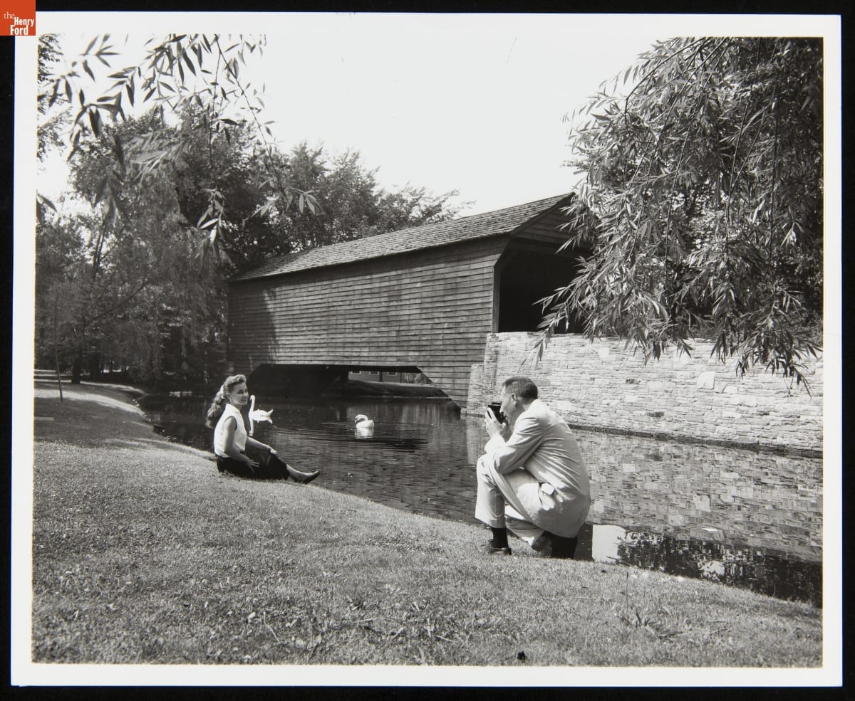 A man, kneeling, photographs a woman sitting on the bank of a pond with a covered bridge in the background
