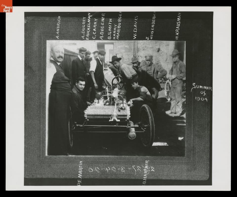 Matted black-and-white photo of group of men around an early open car; names written on the mat