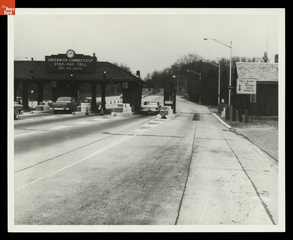 Black-and-white photo of tollbooths with cars stopped at them on wide roadway
