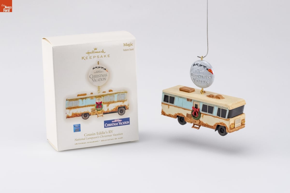 Ornament of rusted beige RV with wreath on door, dangling from round silver button reading "National Lampoon's Christmas Vacation," sitting next to a white box with an image of the same ornament on it