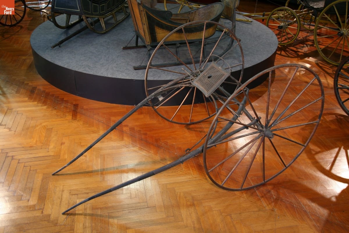 Minimal conveyance with two large wheels, rudimentary seat, and bars for attaching to a horse