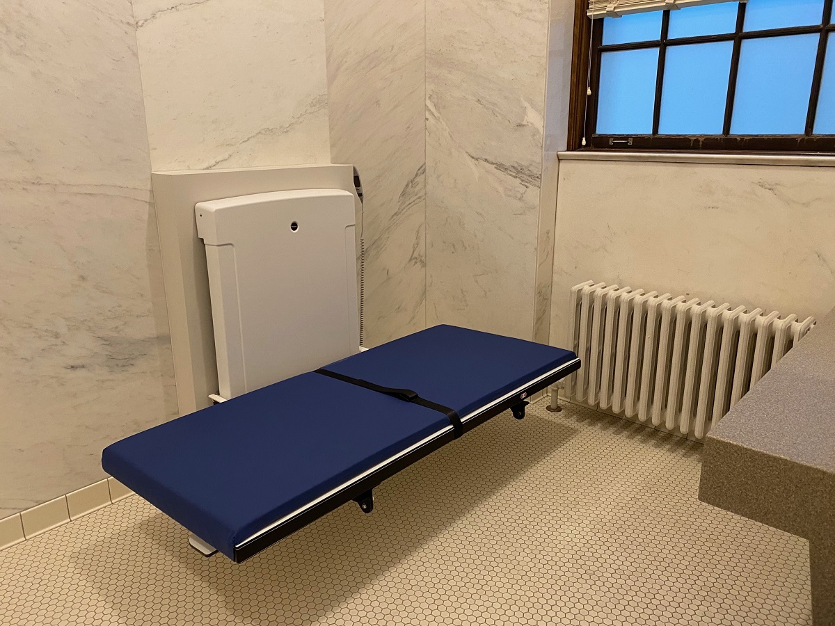 Blue padded table with machinery behind it in a space with tile floor and marble walls (also a window and radiator)