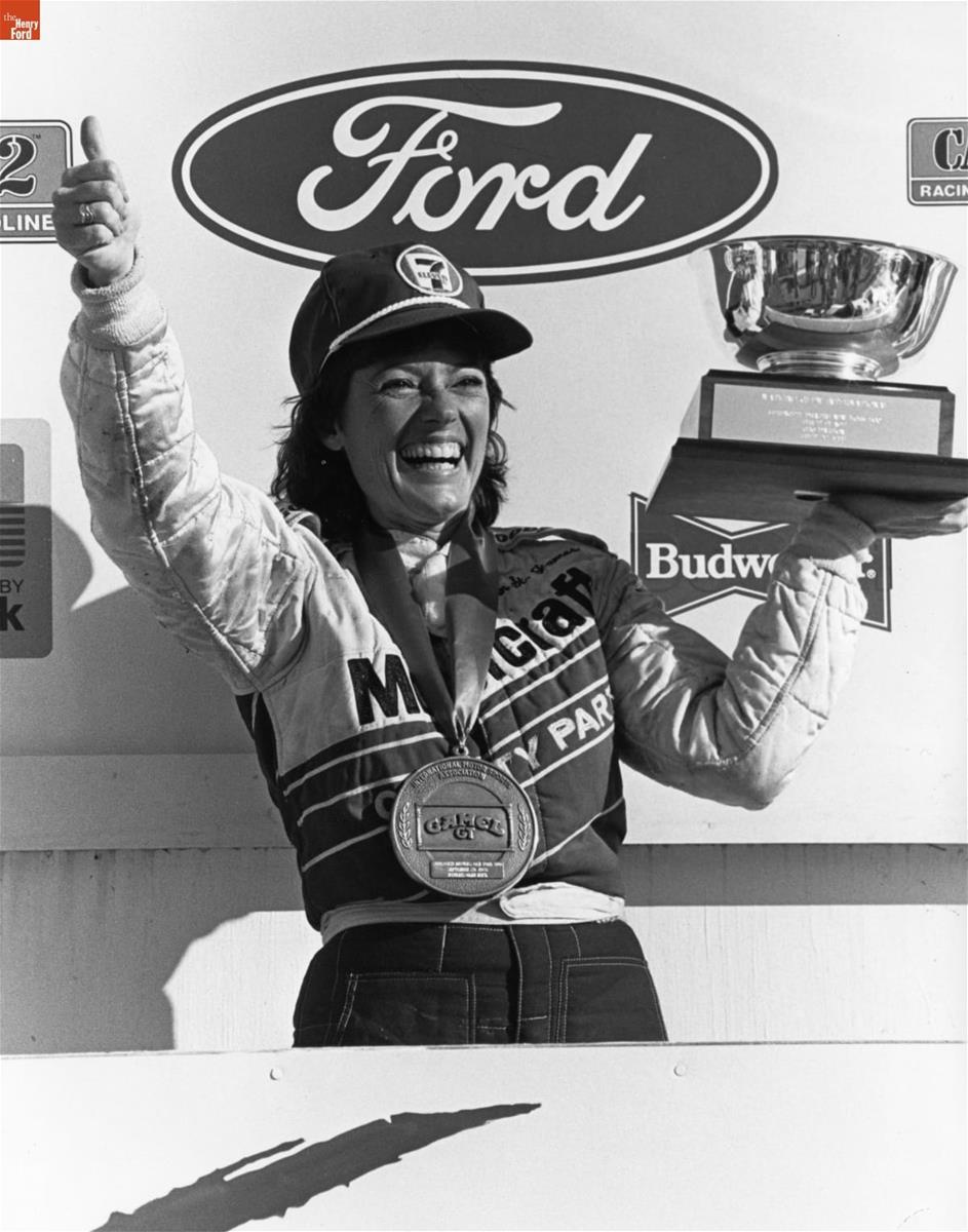 Smiling woman in a jumpsuit and baseball cap, with large medal around her neck, holds a trophy in one hand and makes a thumbs-up with the other, in front of a wall with Ford and Budweiser logos, among others