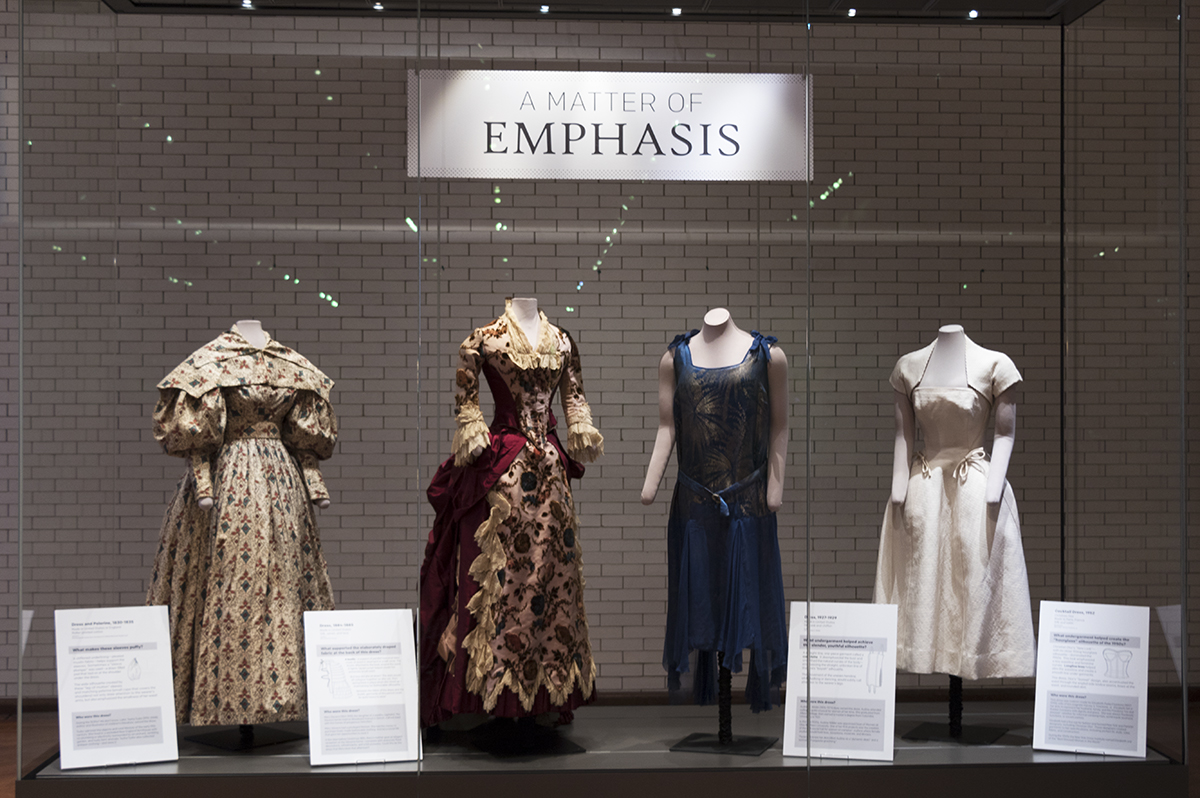 Display case containing four dresses on mannequins, each with a sign nearby
