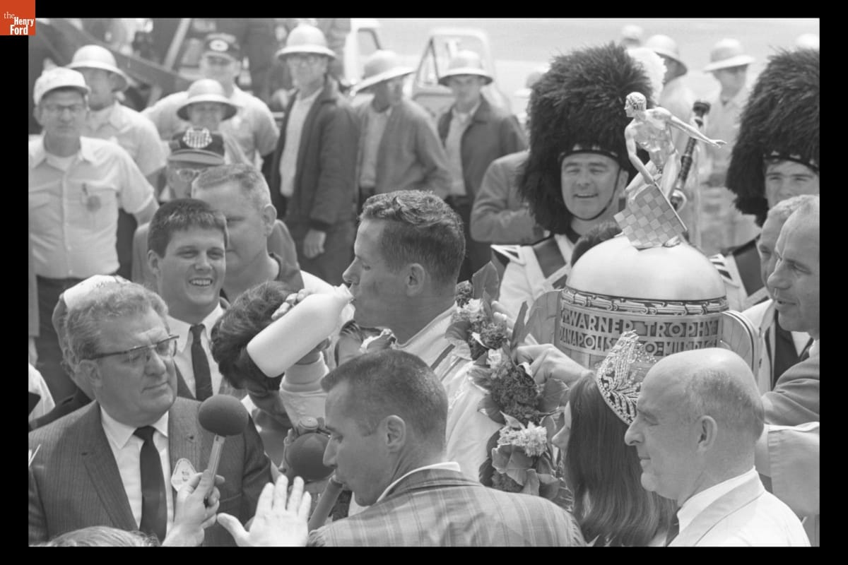 Man drinks from glass milk bottle in the midst of a crowd with a large trophy and men in beefeater hats nearby