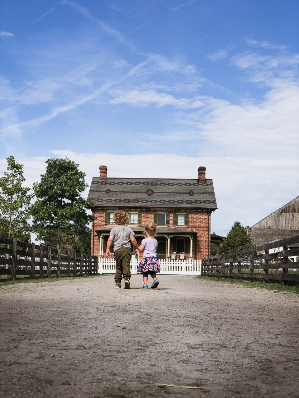 Two children walk toward Firestone Farm in Greenfield Village. Two children walk down gravel lane between wooden fences toward a red brick farmhouse