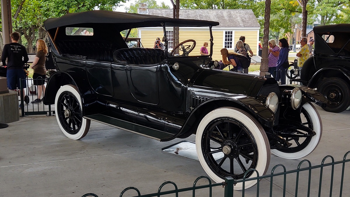 Old-fashioned black car with open sides and dash and whitewall tires, with people standing nearby