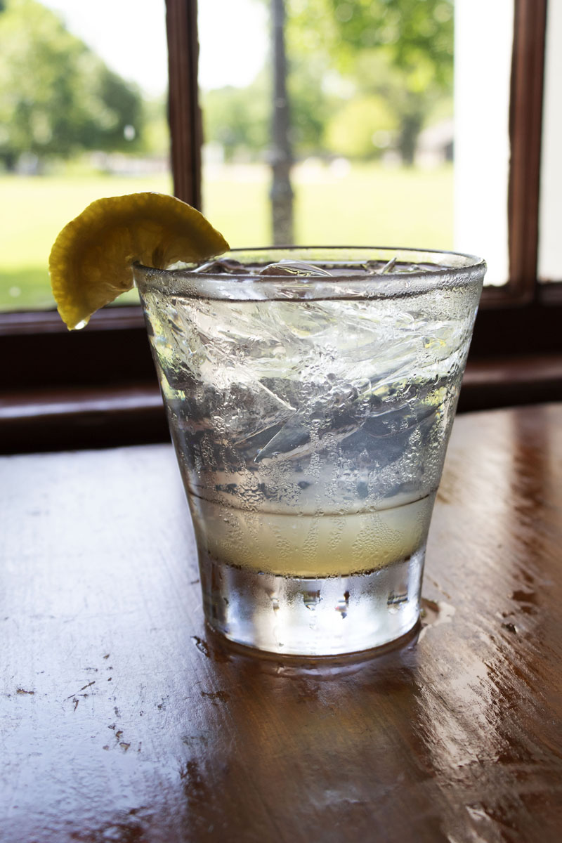 Clear glass with lemon on rim, filled with ice and beverage, sitting on a wooden table with a window in the background