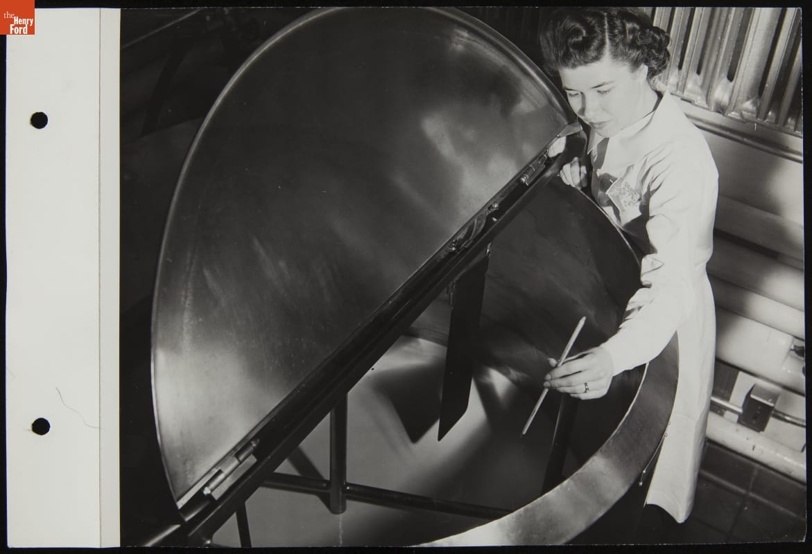 Woman holds stick or gauge over large metal vat containing liquid