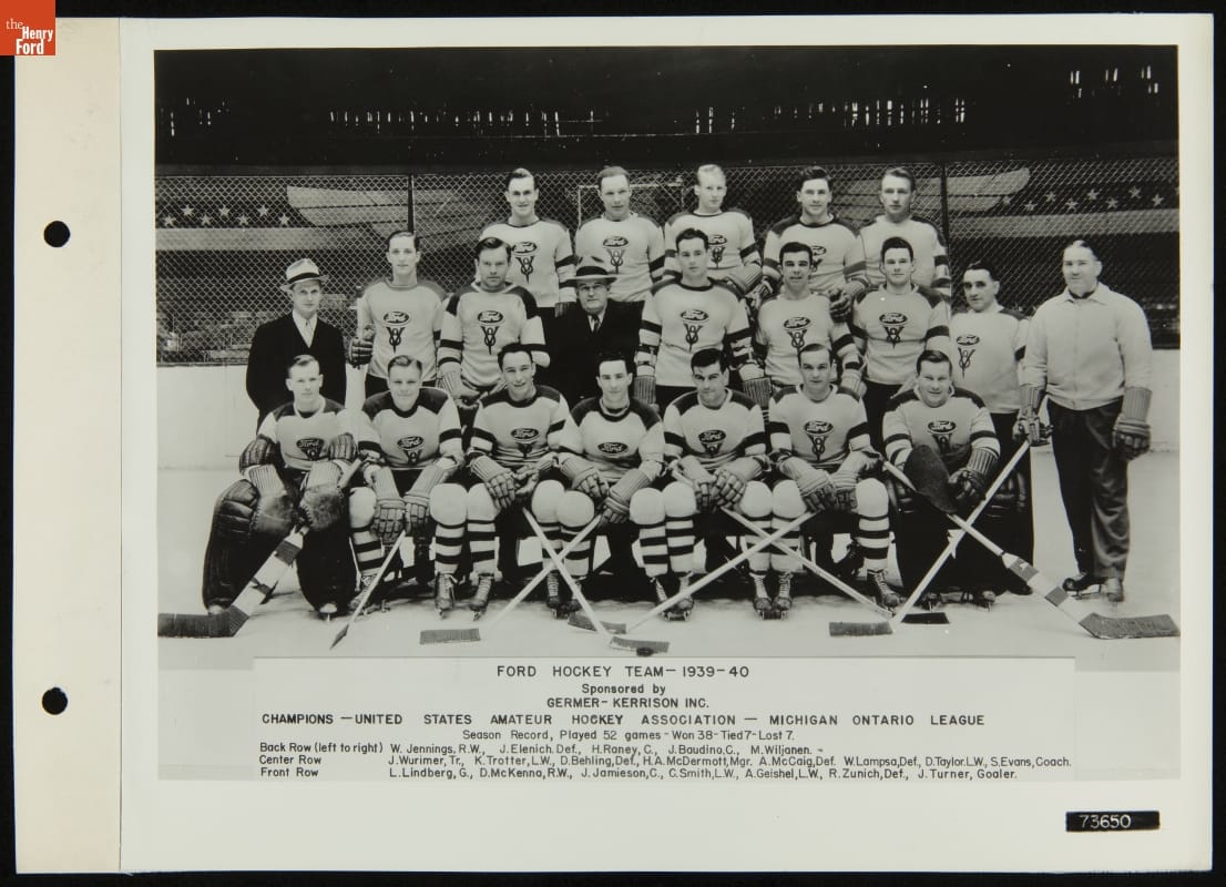Group of men, most in hockey uniforms, some with hockey sticks, pose on ice in what appears to be an indoor ice rink