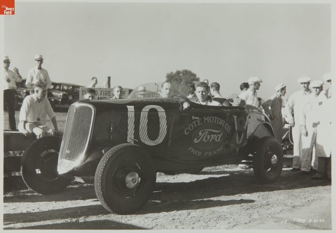 Man sits in early, open racecar with other cars and people in the background