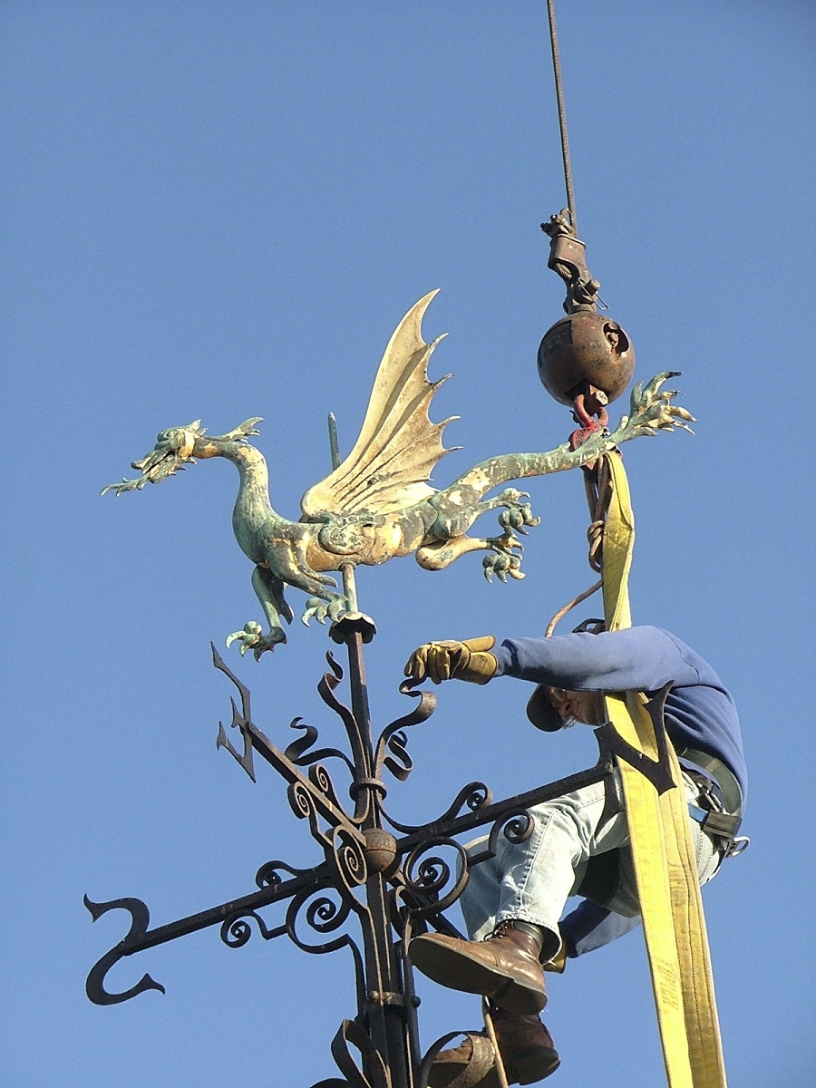 Removing the dragon weathervane Man suspended from crane holds onto an elaborate metal weathervane in the shape of a dragon with decorative elements underneath