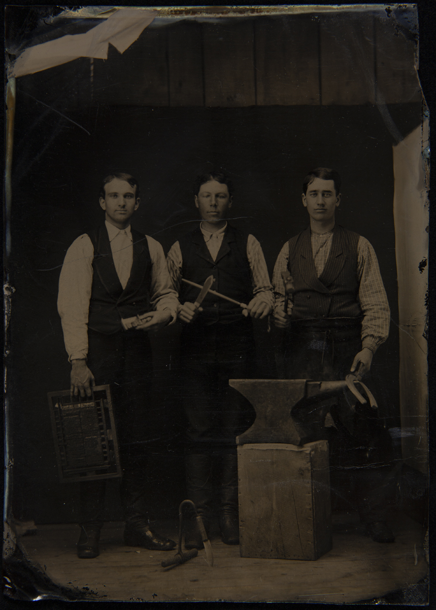 Three men in vests and shirtsleeves standing behind an anvil on a wooden block, holding tools