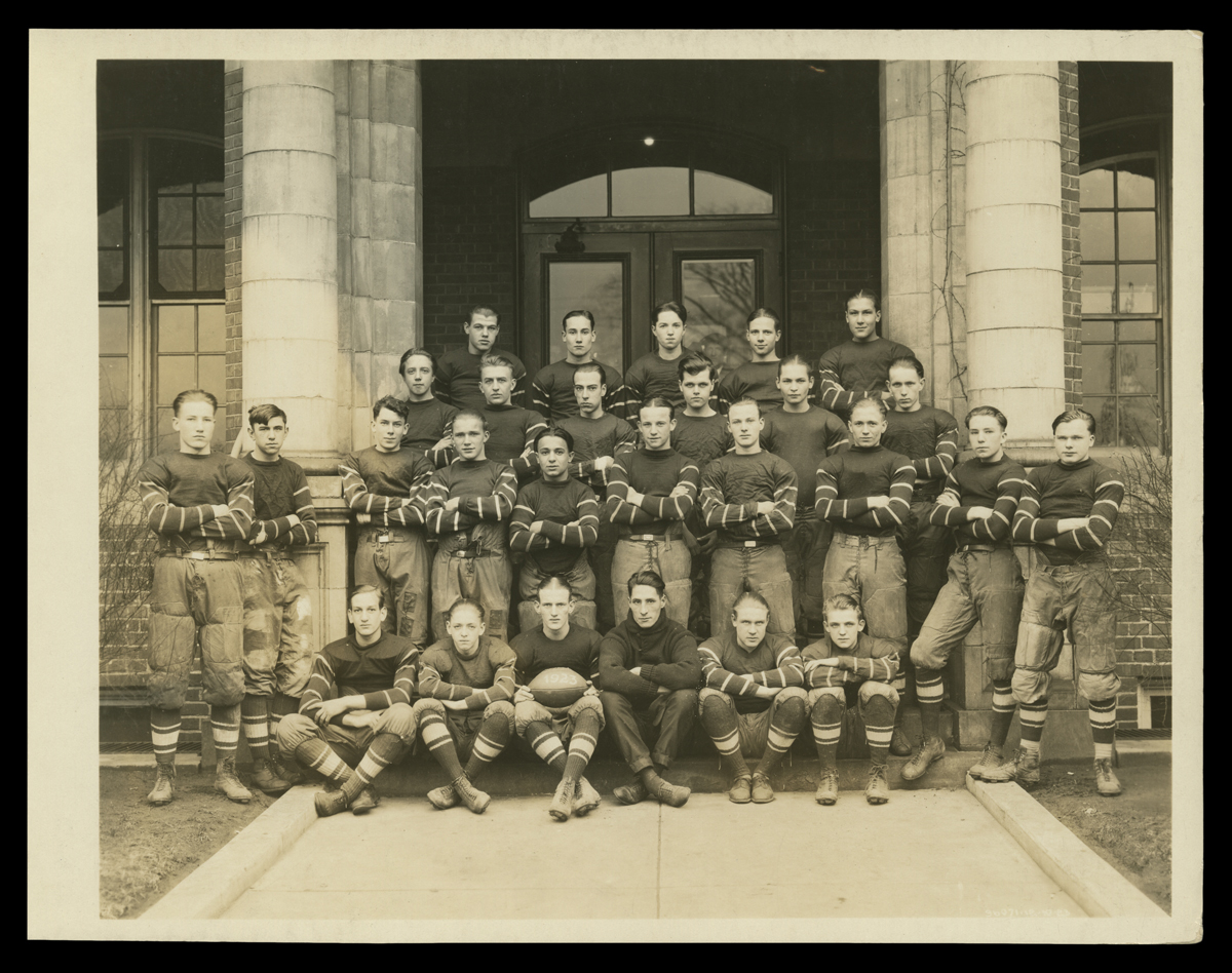 Group of boys/young men wearing sports uniforms pose with arms crossed, seated and standing, in front of a building 