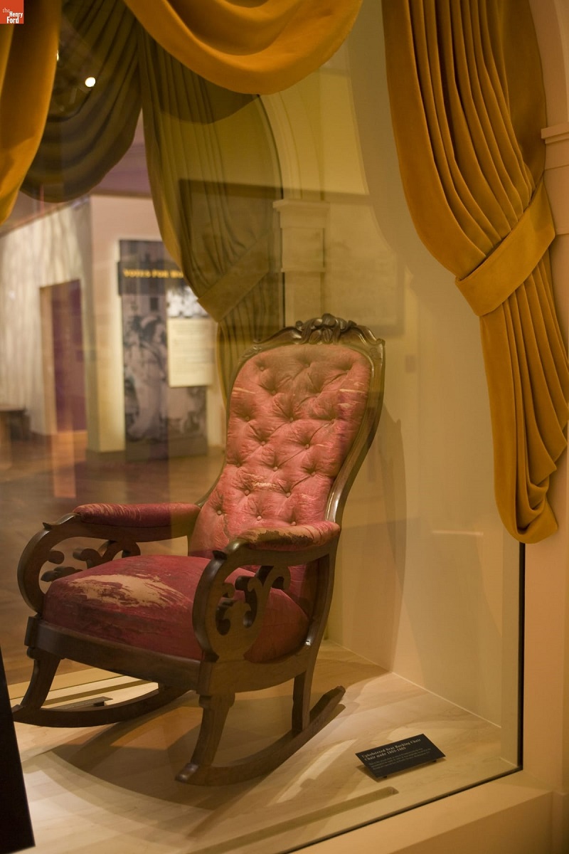 Red upholstered rocking chair in glass case surrounded by mustard yellow curtains