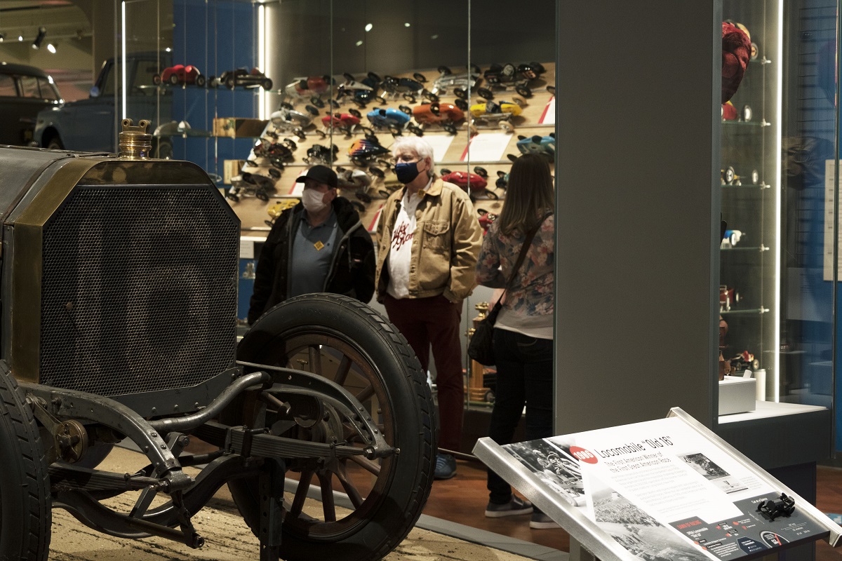 Several people look at a museum exhibit, standing among display cases and cars