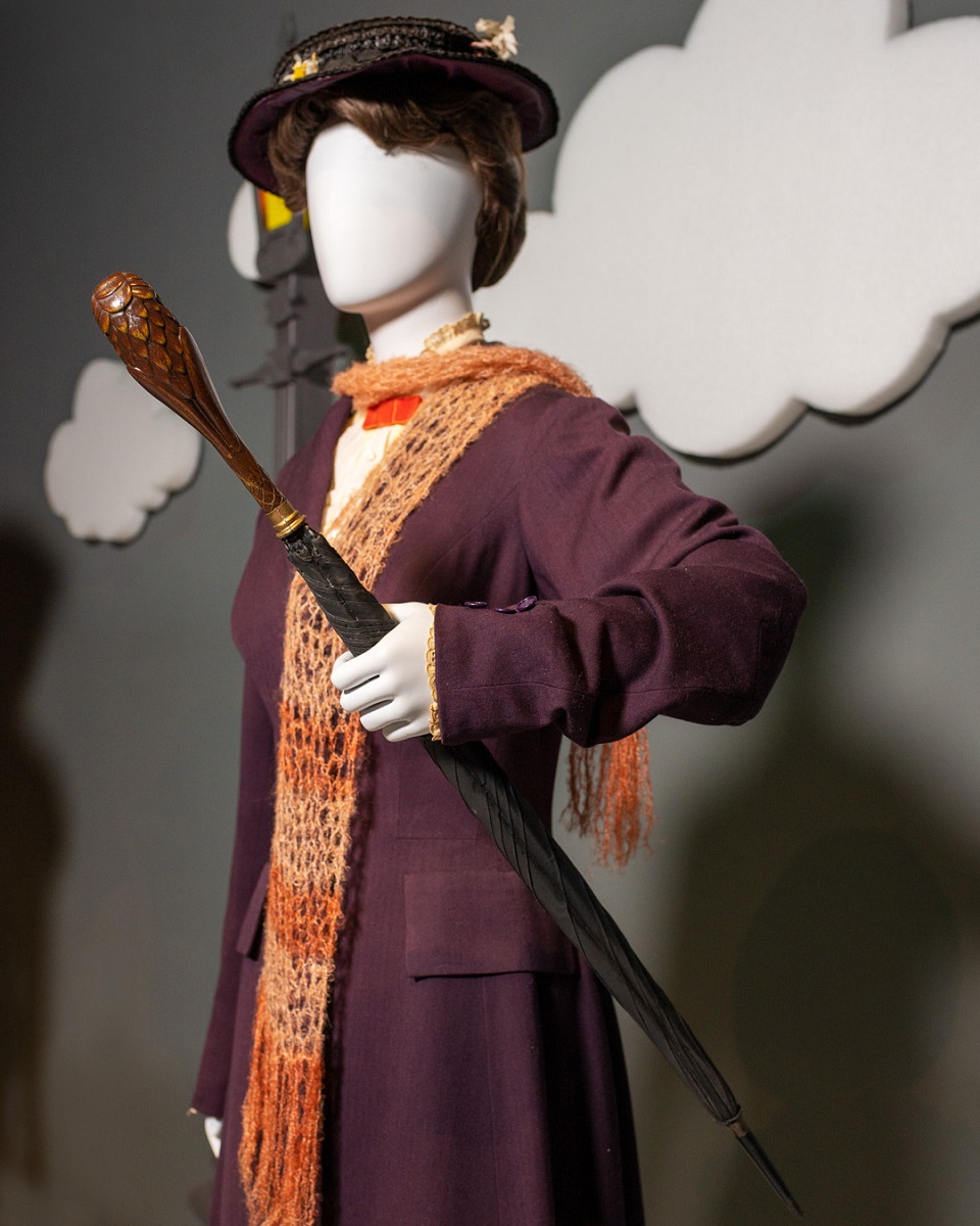 Mannequin dressed in a dark purple coat with an orange knitted scarf, black hat, and umbrella with carved wooden handle, posed in front of a gray wall with white clouds