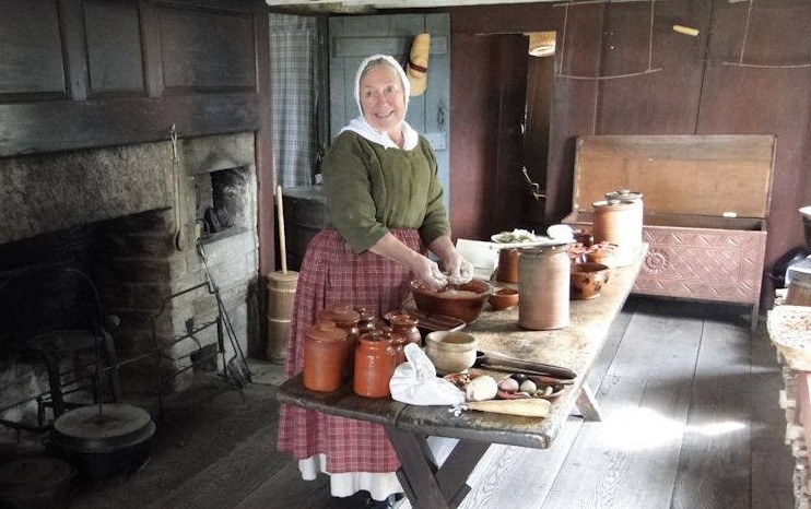 Woman works at a rustic table in a kitchen with wooden walls and large rustic fireplace