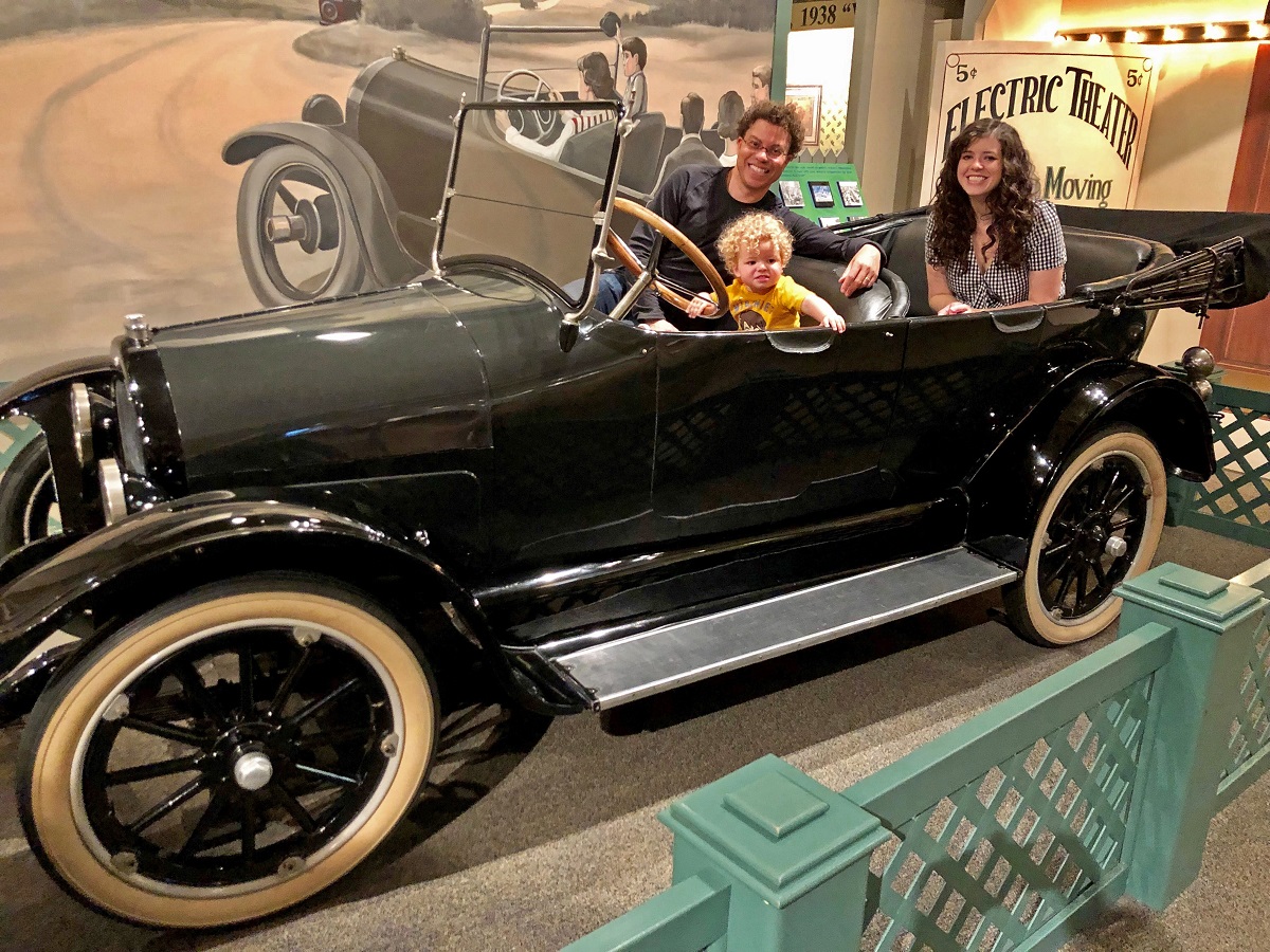A man, woman, and child sit in a convertible car with a backdrop and other items in the background