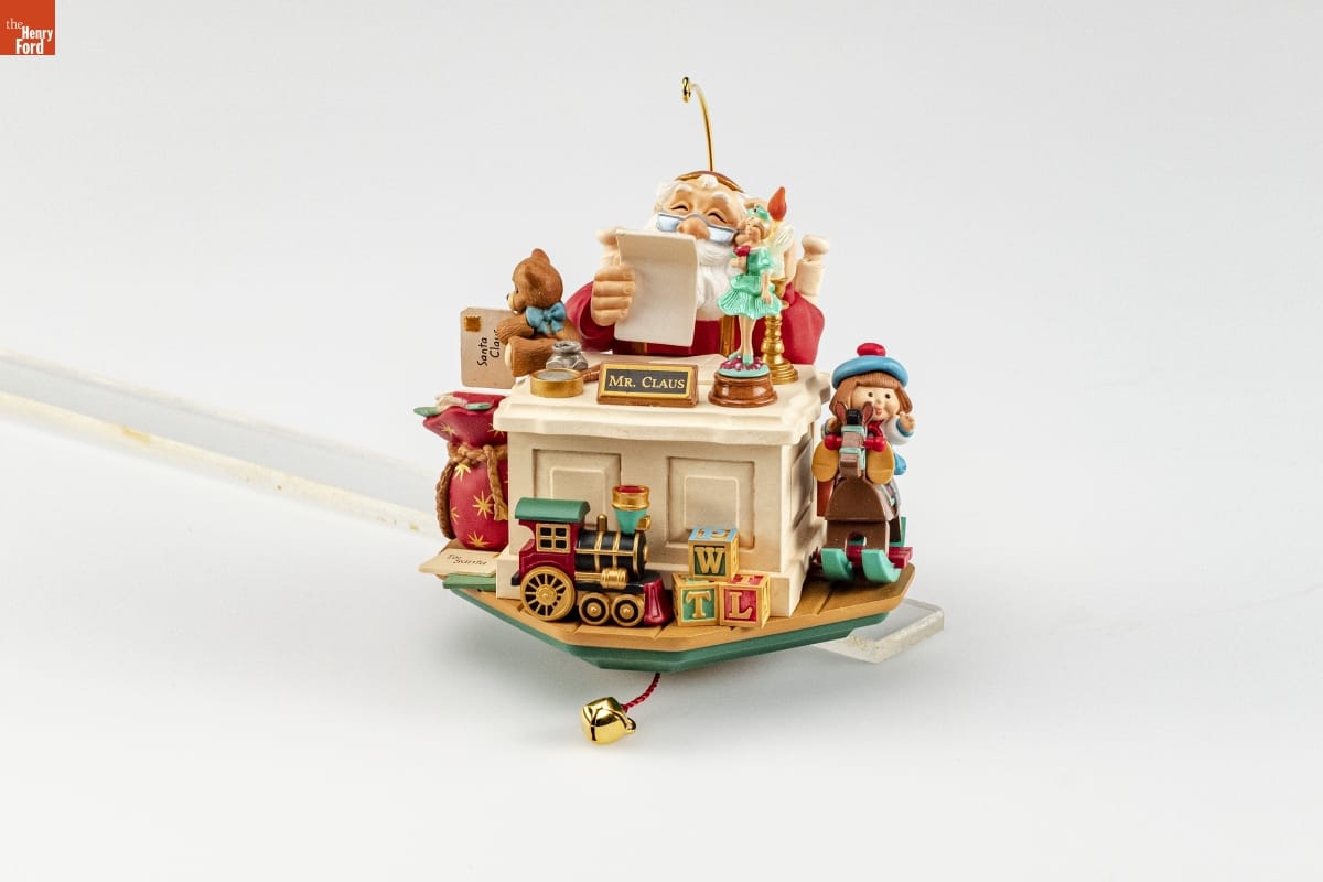Ornament depicting Santa Claus sitting behind a desk with sign "Mr. Claus," looking at a piece of paper, with many toys on and around the desk