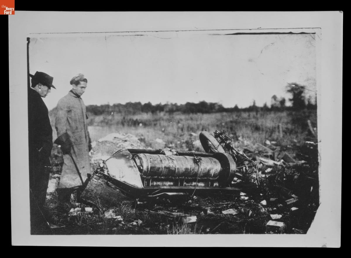 Black-and-white photo of two men staring at the crashed remains of a car in a field