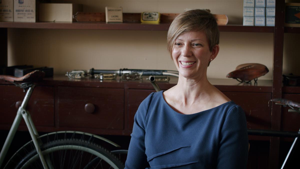 Woman in blue top smiling, bicycle and counter/shelves partially visible behind her