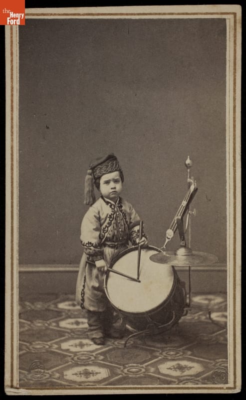 Small child in elaborate costume plays a large drum with drumsticks
