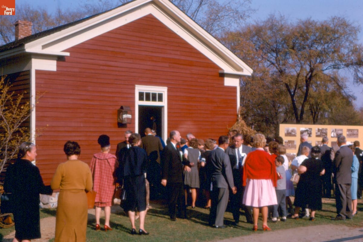 Group of people mingle outside a small, red, wooden building