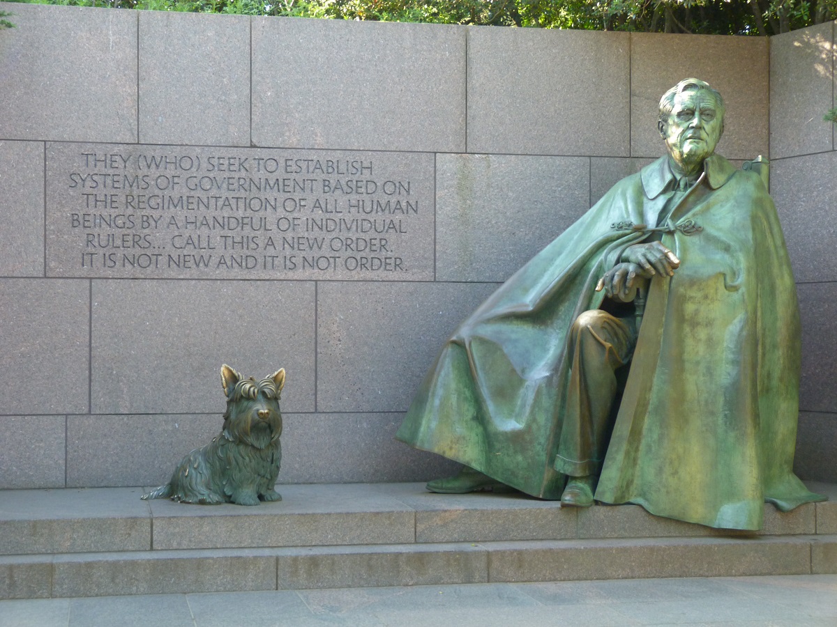 Statue of a seated man wearing a cape next to statue of a Scottie dog, mounted on granite with a granite wall with inscribed text behind them