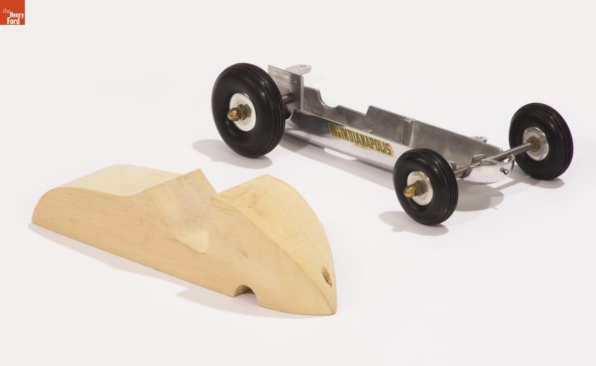 Metal model car chassis sitting next to carved, unpainted block of wood in the shape of a car body