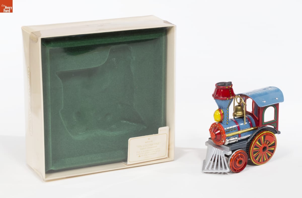 Colorful ornament of a blue steam locomotive sits next to a cream-colored box with green velvet insert and clear plastic top