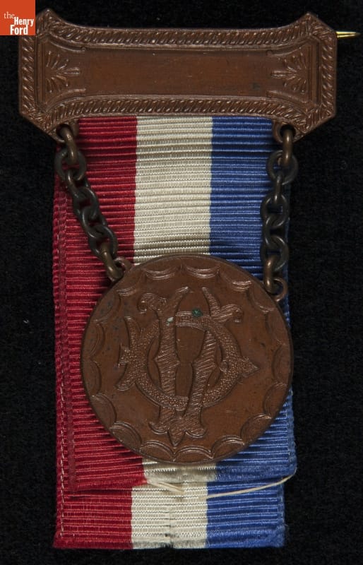 Metal (?) and fabric badge with red, white, and blue stripes and intertwined letters