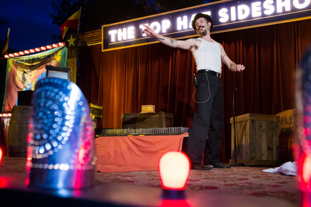 The Top Hat Side Show performing at Hallowe’en in Greenfield Village for the first time in 2015 Man in black pants and hat and white tank top points from a stage, with lighted "Top Hat Side Show" sign behind him