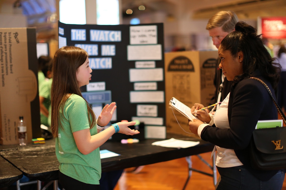 A young girl in a green t-shirt talks to two other people in front of a trifold display board on a table