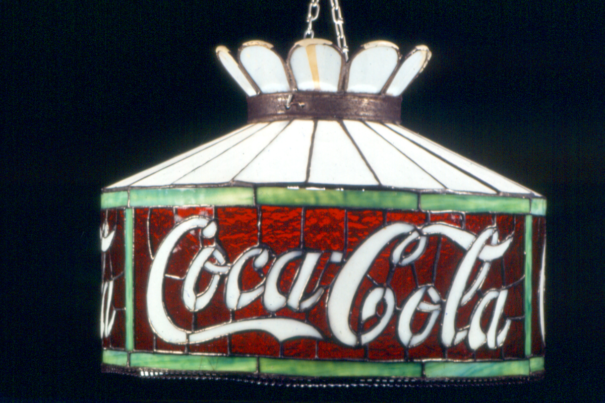 Stained glass lamp in shades of white, green, and red, with white cursive text "Coca-Cola"