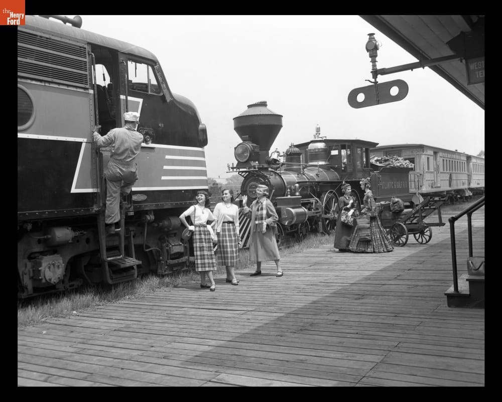 Two locomotives face each other on a track, with women in period clothing on the platform in front of them