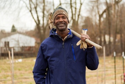 Smiling African American man in blue jacket and knit hat holding shovel on shoulder