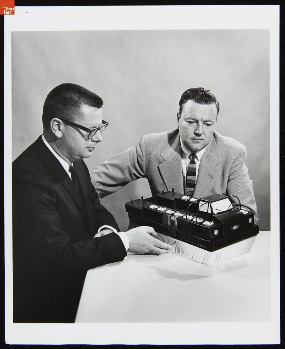 Two men in suits sit at a table, looking at a model of a truck-like vehicle with white fringe below body