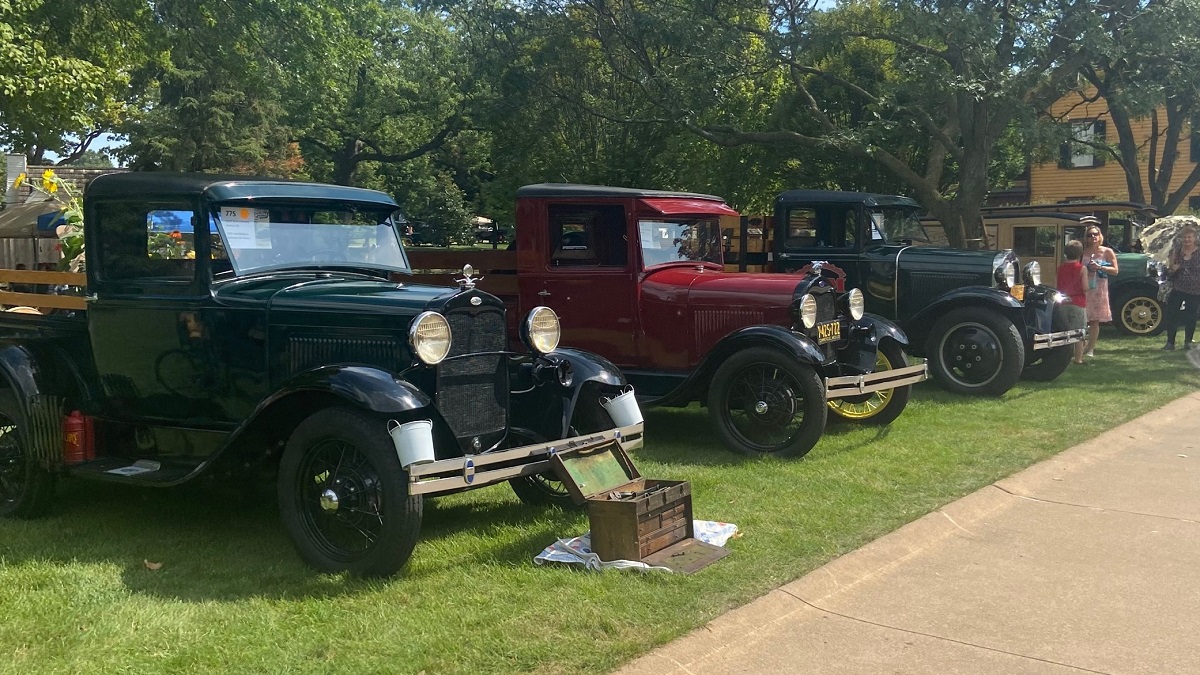Old-fashioned trucks are parked in a line on a green lawn with large trees and buildings behind them and people nearby