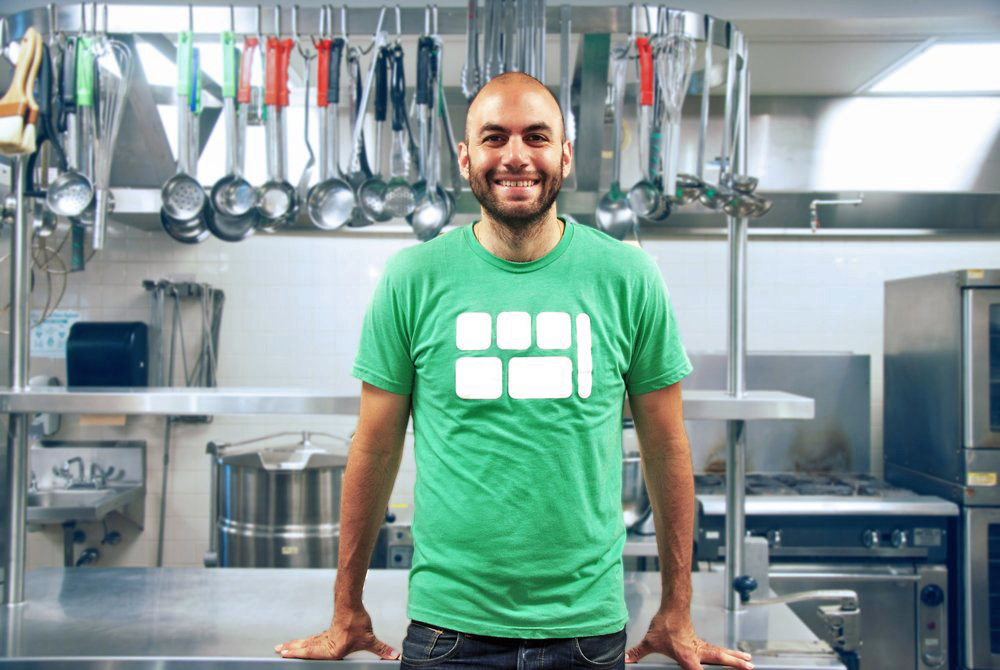 Man in green t-shirt with white graphic leans against a stainless steel table in an industrial kitchen