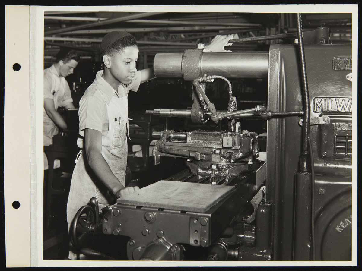 African American boy wearing apron and skullcap works at machine; another boy at another machine in background