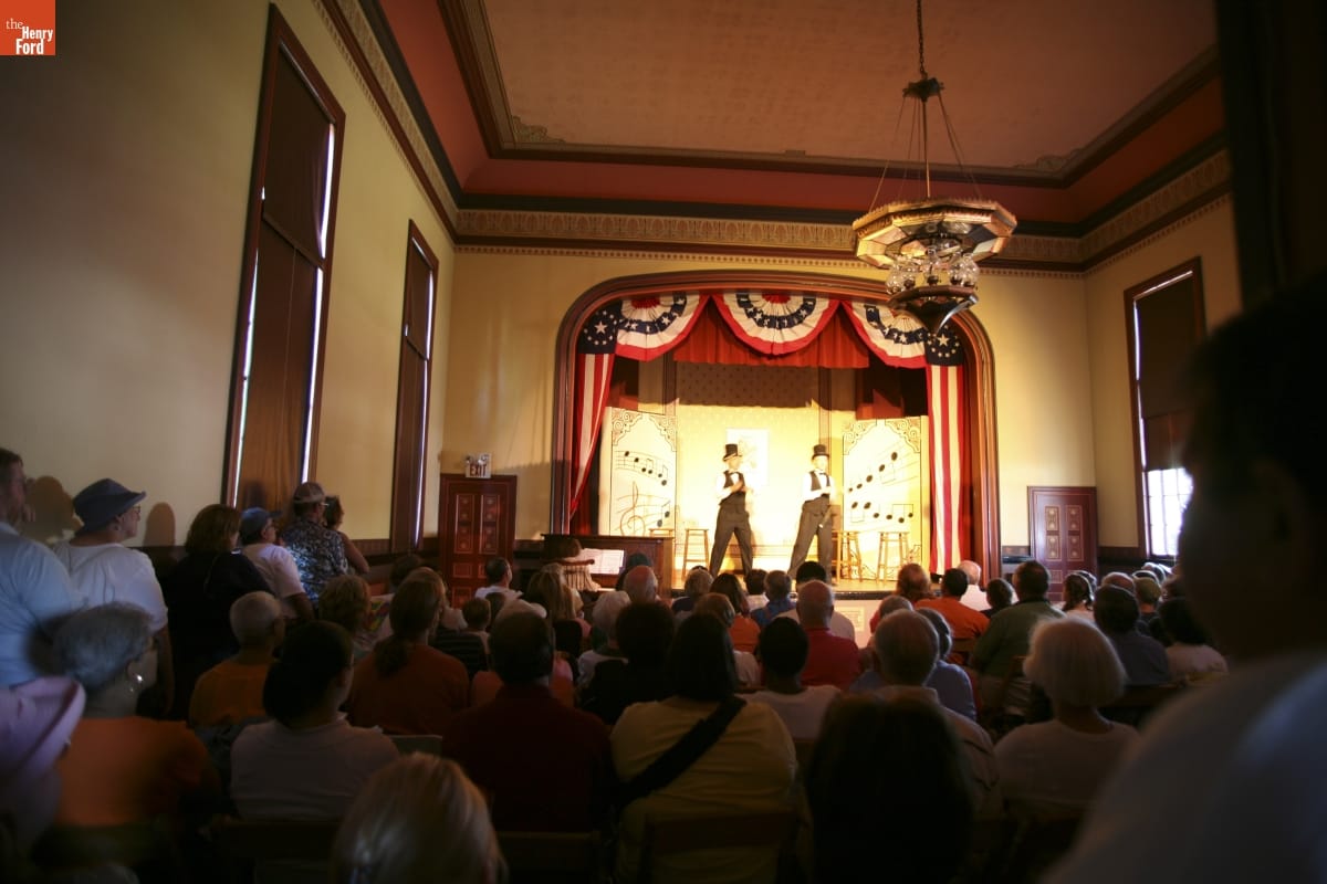 Two people on a stage surrounded by red, white, and blue bunting perform in an auditorium filled with people