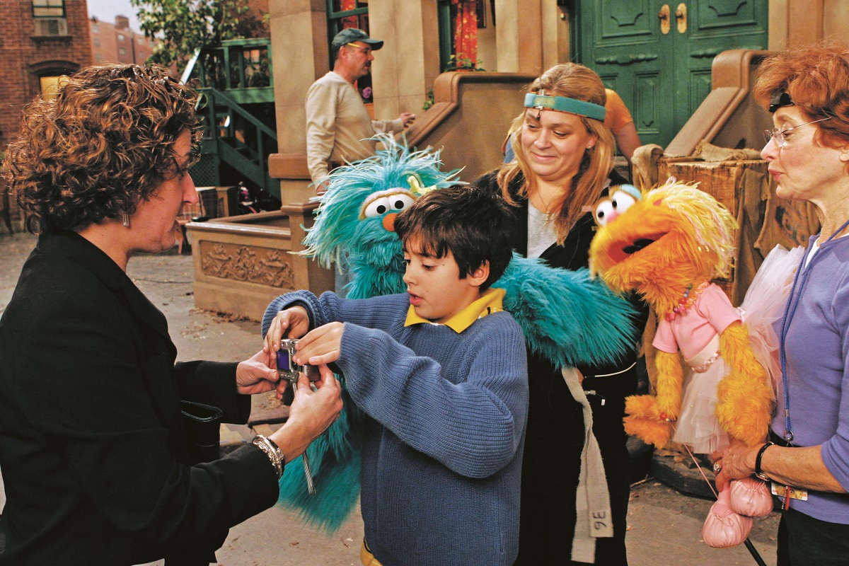 Group of people, two holding furry puppets, on TV set designed to look like a street